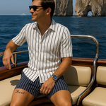 Ein Mann in einem blau-weiß gestreiften Sommer-Set, bestehend aus Kurzarmhemd und Shorts, sitzt auf einem Holzboot vor den Faraglioni-Felsen auf Capri.