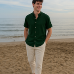 Ein lächelnder Mann in einem dunkelgrünen Kurzarmhemd mit Stehkragen und einer hellen Hose spaziert am Sandstrand entlang, mit dem Meer im Hintergrund.