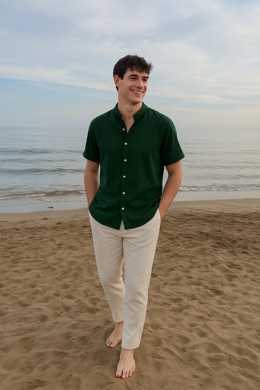 Ein lächelnder Mann in einem dunkelgrünen Kurzarmhemd mit Stehkragen und einer hellen Hose spaziert am Sandstrand entlang, mit dem Meer im Hintergrund.