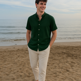 Ein lächelnder Mann in einem dunkelgrünen Kurzarmhemd mit Stehkragen und einer hellen Hose spaziert am Sandstrand entlang, mit dem Meer im Hintergrund.