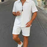 Ein Mann in einem weißen Sommer-Set mit einer 3D-Waffelstruktur, bestehend aus einem Kurzarmhemd und passenden Shorts, geht eine Straße entlang.

